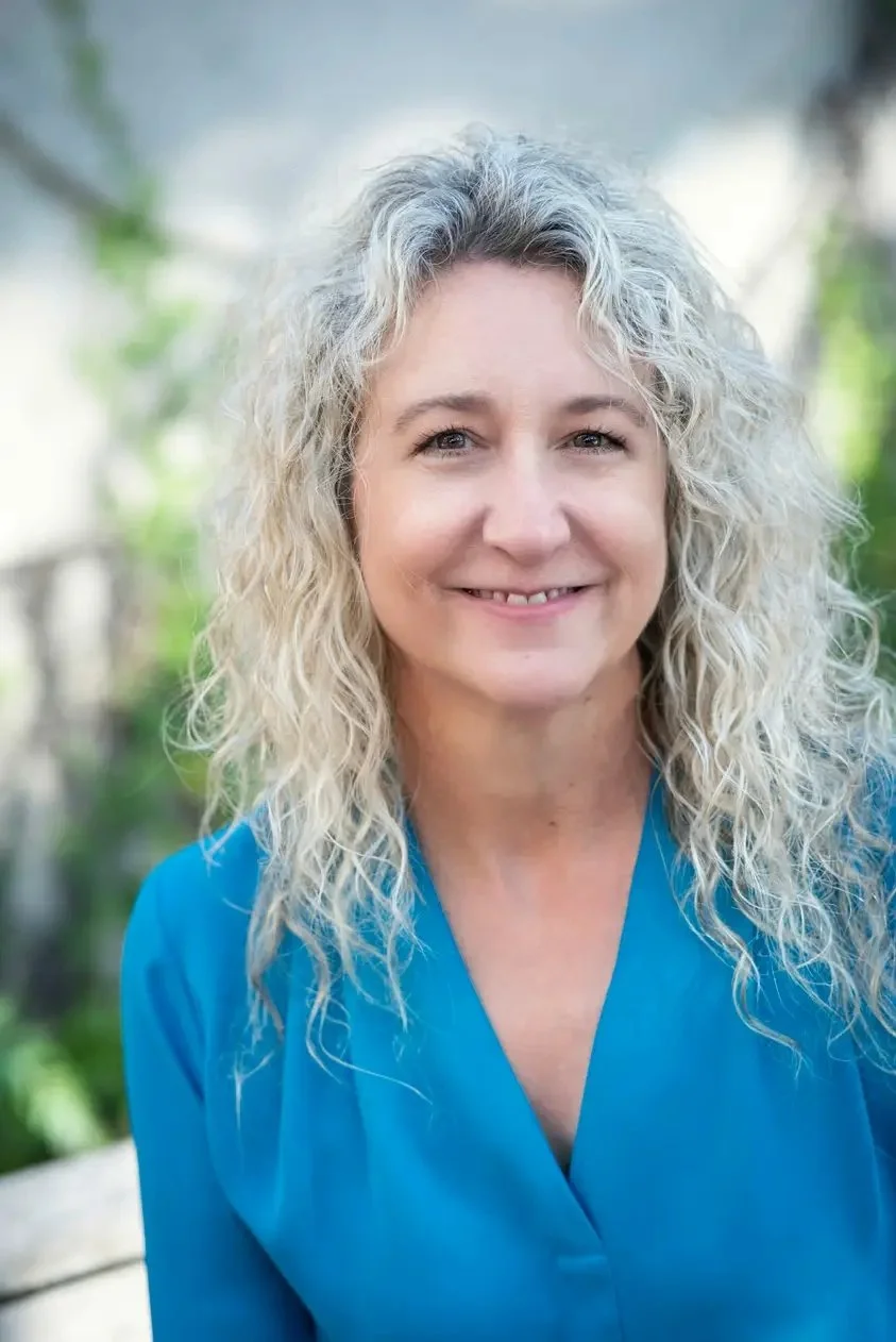 Professional headshot of Nancy Benet smiling, with curly blonde hair and a bright blue blouse, photographed outdoors against a softly blurred green background.