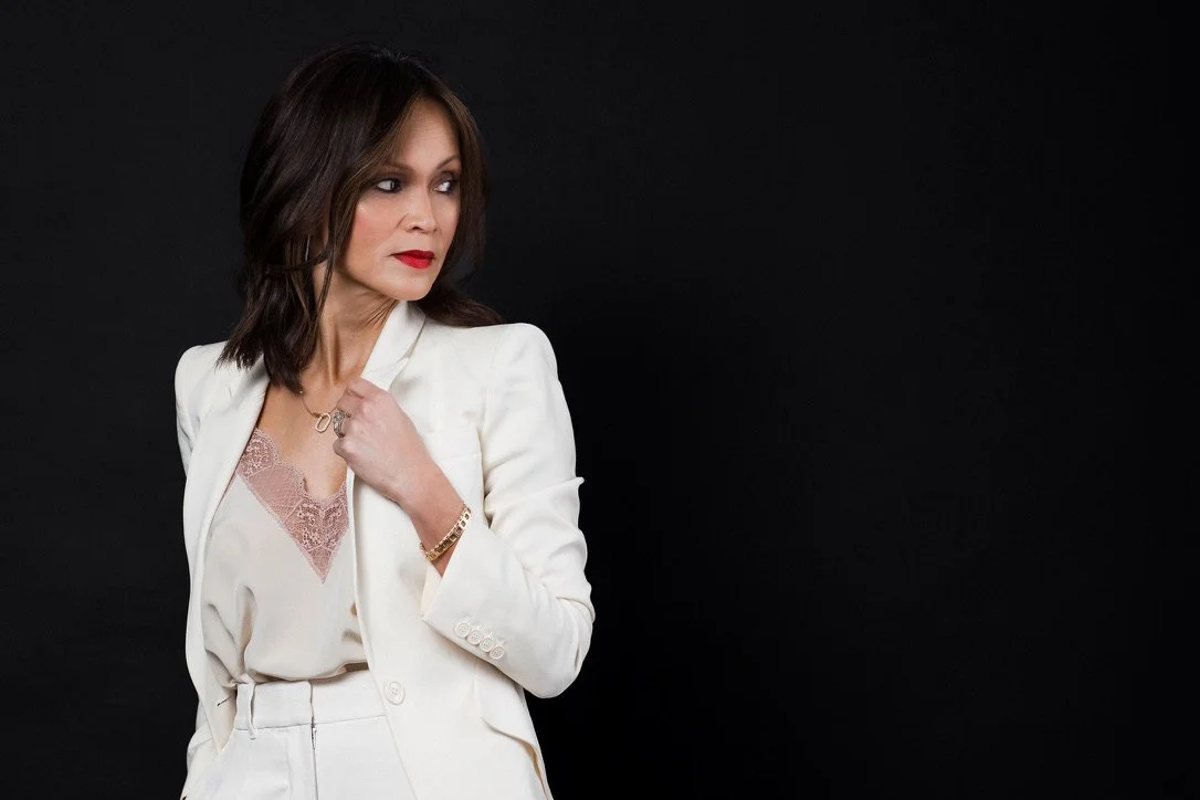 Professional portrait of Soon Hagerty wearing a cream white tailored suit with a lace camisole, looking confidently to the side against a dramatic black background. The image reflects strength, sophistication, and modern female leadership.