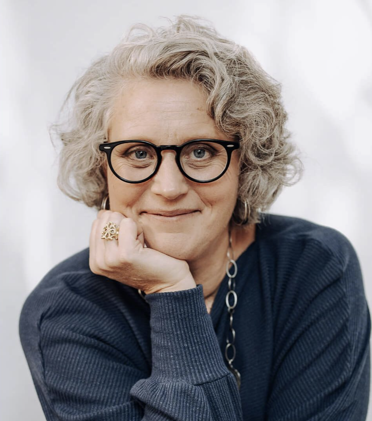 Portrait of Dr. Laura James, a woman with short gray curly hair and black round glasses, resting her chin on her hand and smiling gently at the camera, wearing a dark sweater and simple jewelry against a light background.