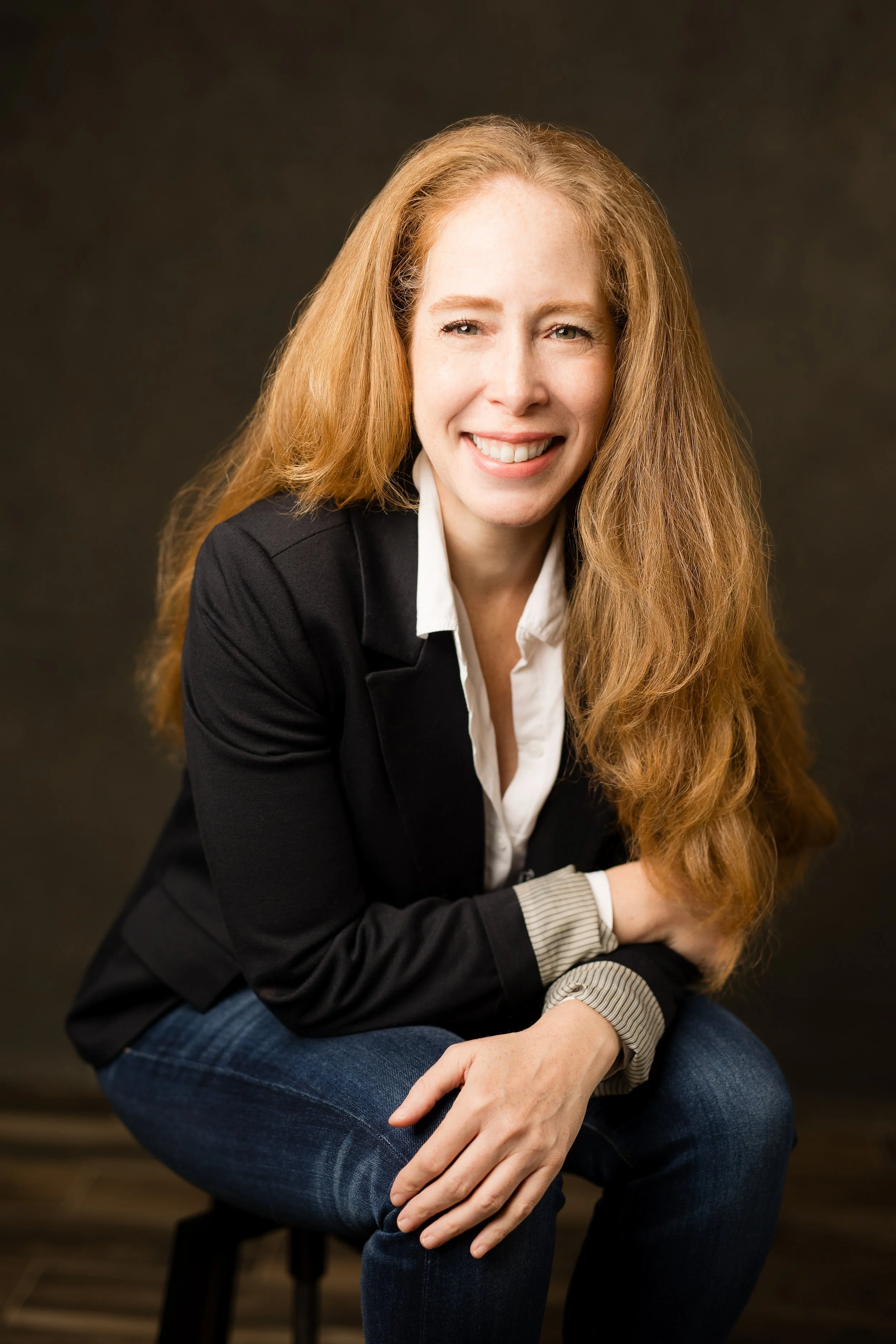 Jane Bertch smiles warmly while seated on a stool, wearing a black blazer, white shirt, and jeans against a dark studio backdrop.