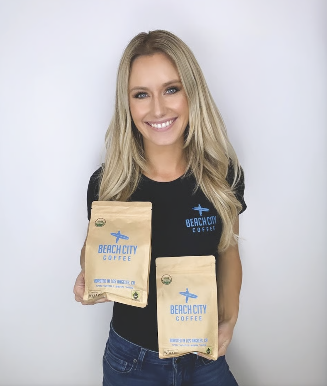 Jillian Salinas, founder of Beach City Coffee, smiles while holding two bags of Beach City Coffee against a light studio background.
