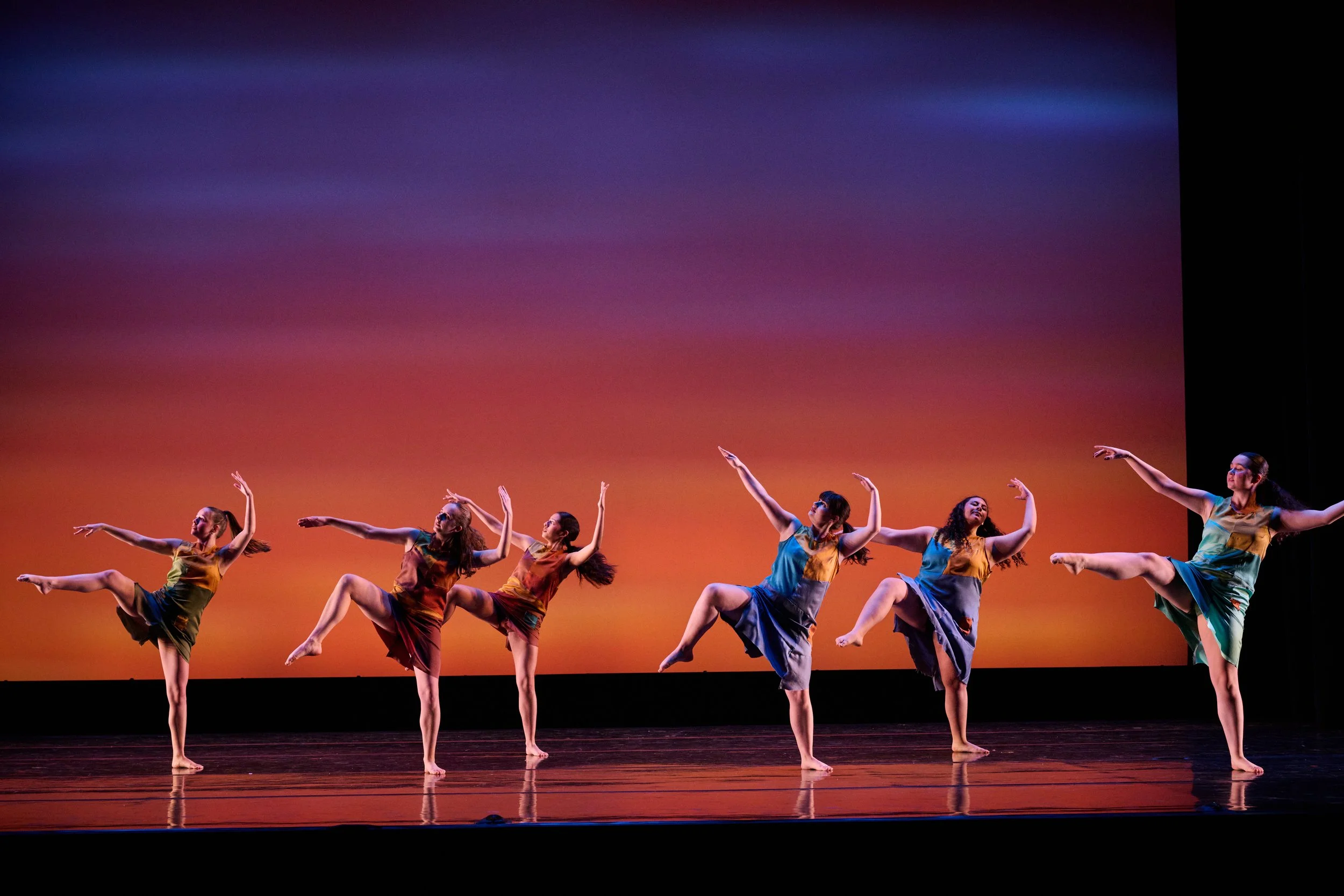 Six dancers on a stage each balancing on one leg with a sunset background