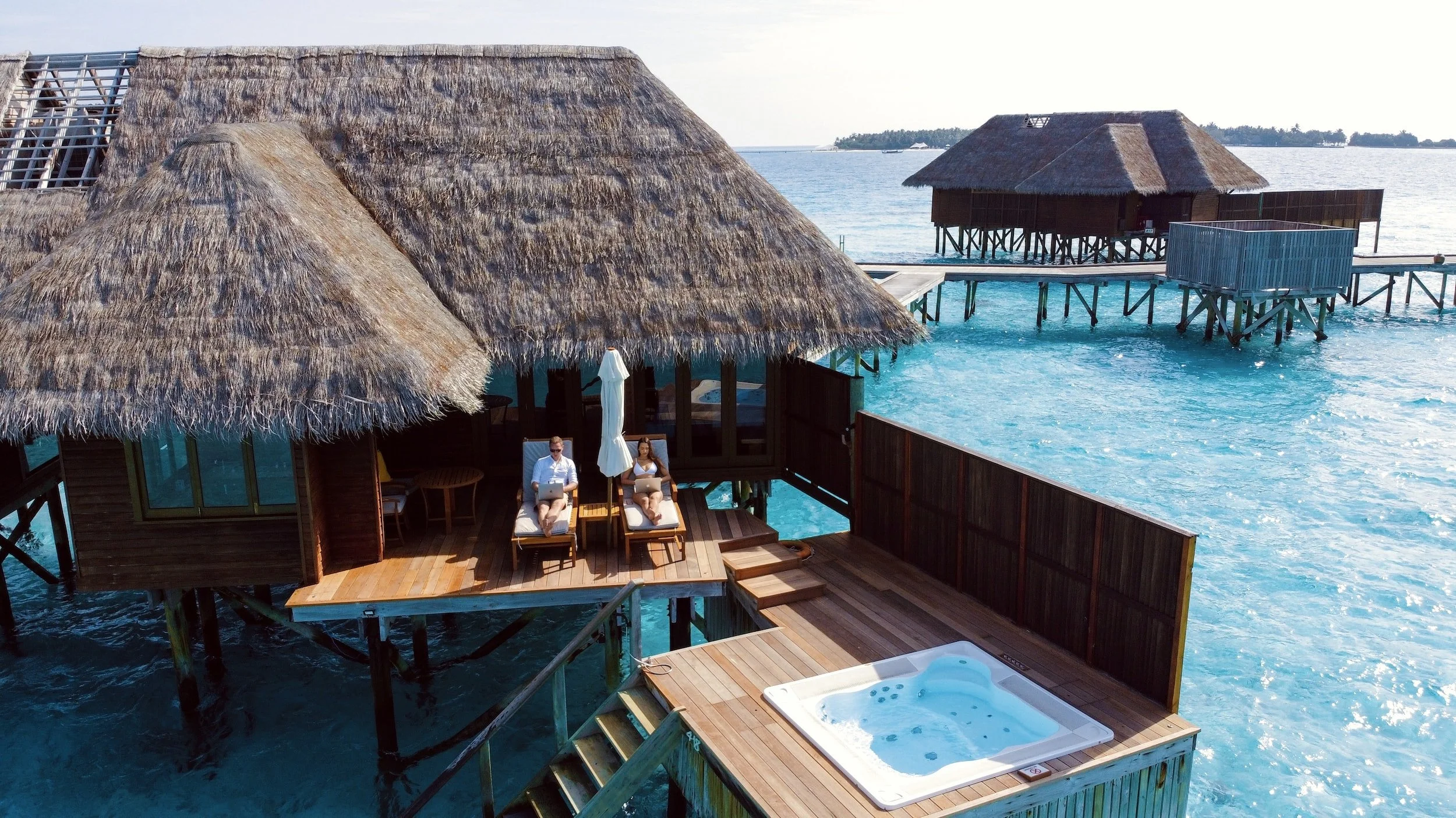 Overwater bungalow with thatched roof, featuring a deck with a hot tub, two sun loungers with people relaxing, surrounded by turquoise ocean.