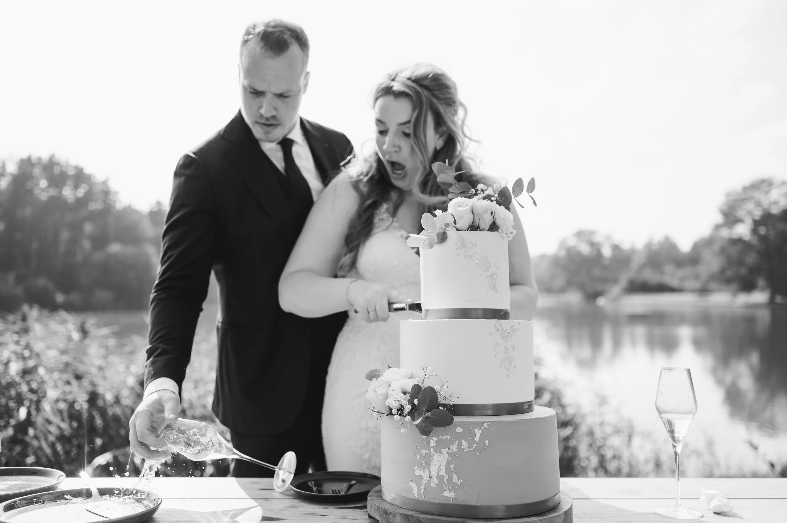 Bruidspaar snijdt de bruidstaart aan bij het water, zwart wit trouwfoto van een echt moment tijdens de bruiloft