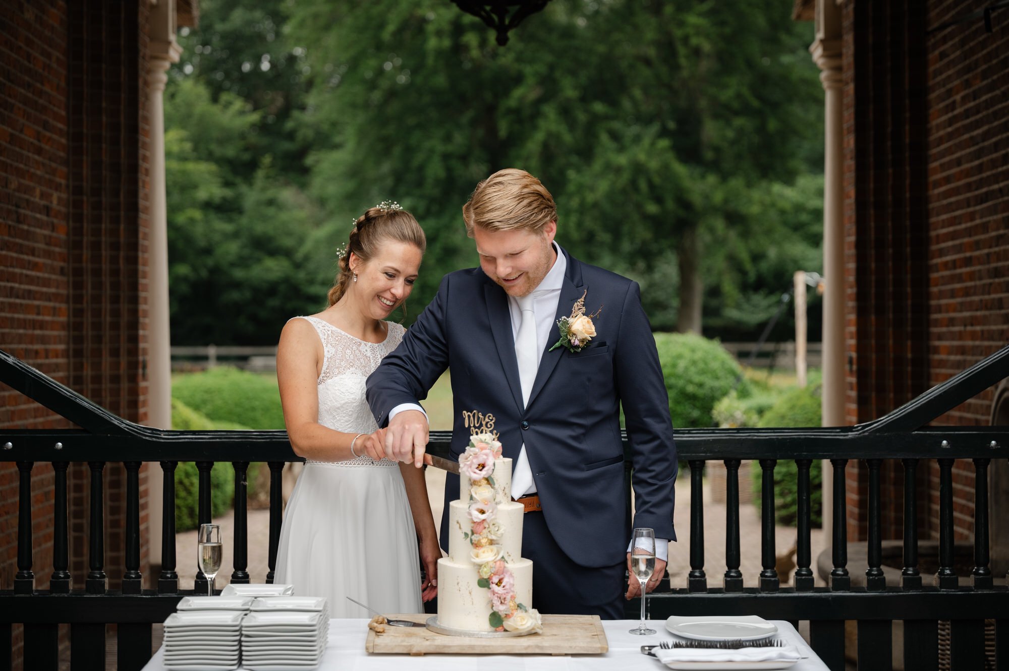 Bruid en bruidegom snijden in een trouwtaart buiten op een terras, met glazen champagne en gedekte tafel ernaast.