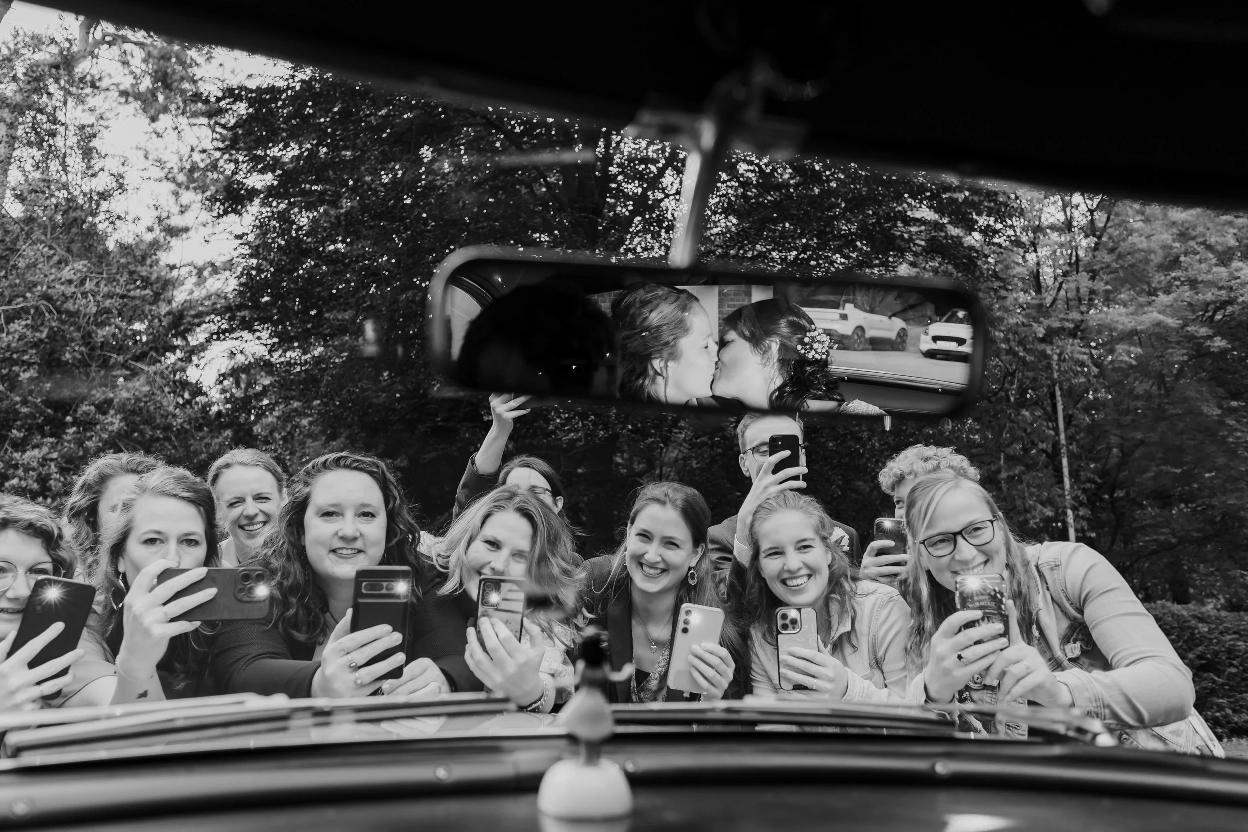   Vriendinnen maken foto’s van Evelien en Monique, met hun kus weerspiegeld in de achteruitkijkspiegel tijdens hun bruiloft bij Villa Klein Heumen  