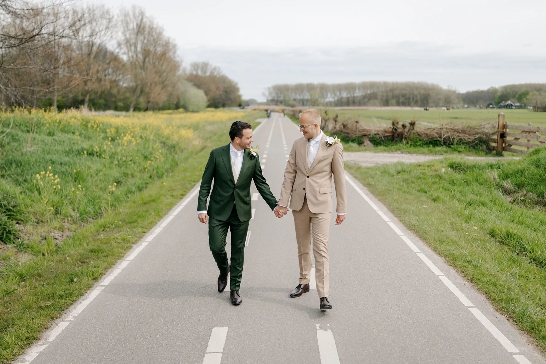  Bruidegoms lopen hand in hand over een landweg tussen bloeiende weilanden 