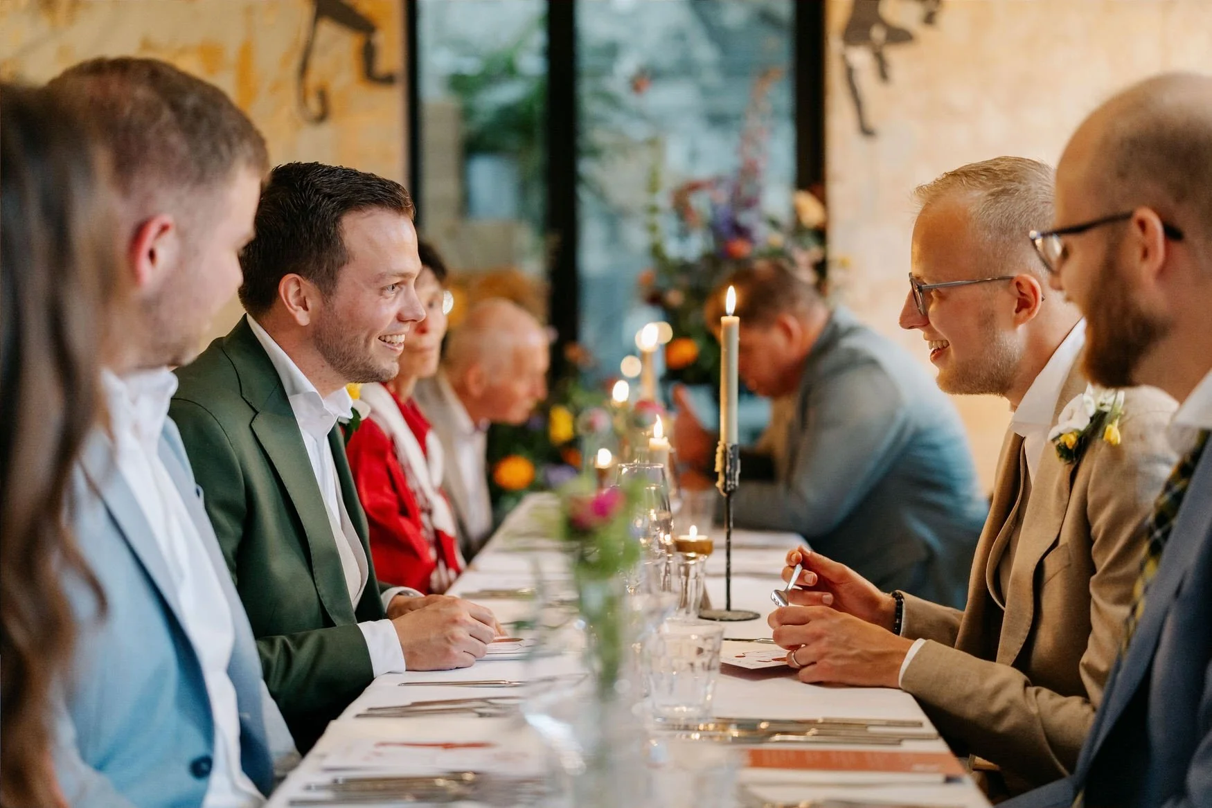  Gasten en bruidspaar aan lange tafel luisteren geamuseerd naar elkaar 
