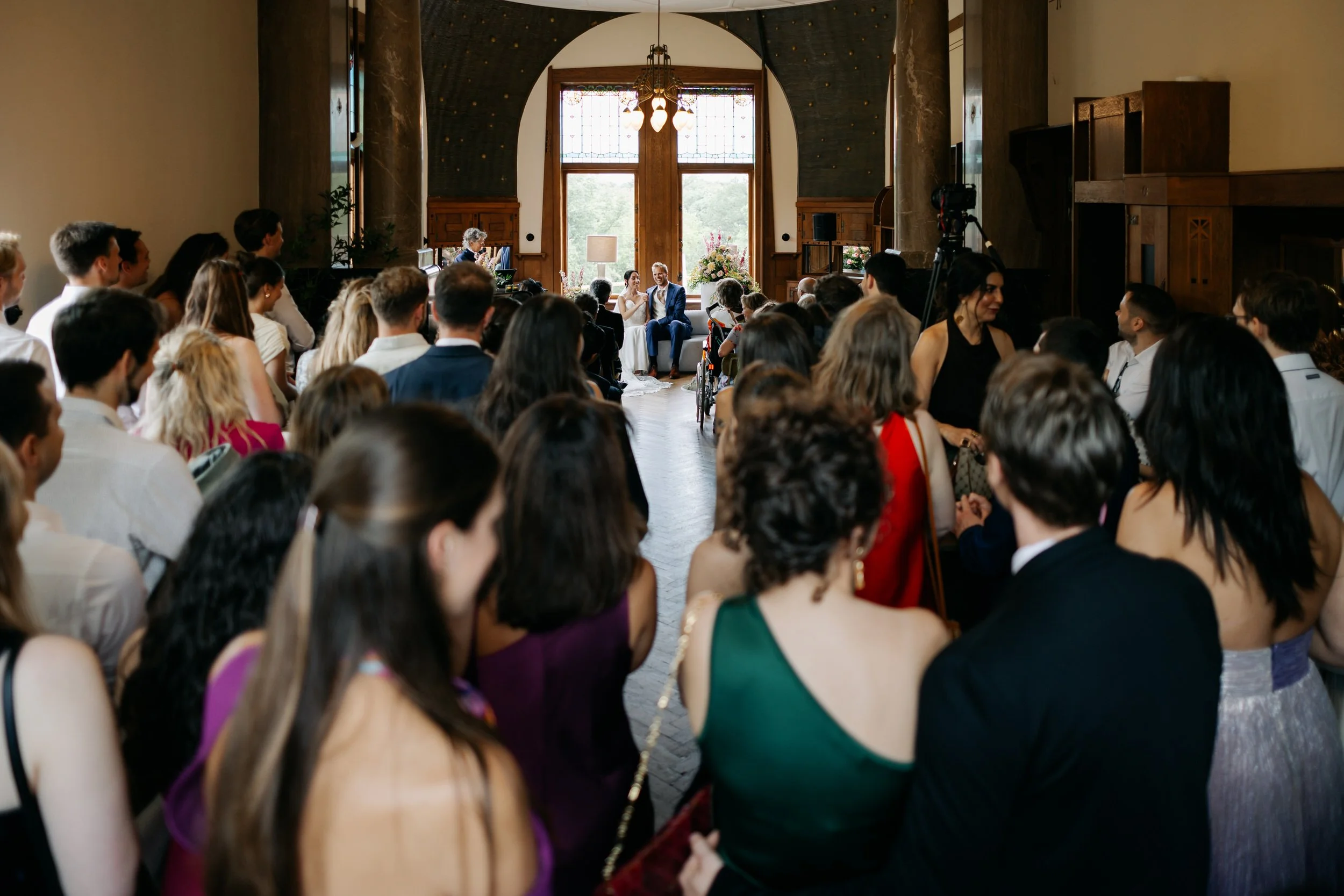  Gasten kijken toe tijdens de ceremonie, gezien vanaf de achterkant van de zaal met de hoge deuren op de achtergrond. 