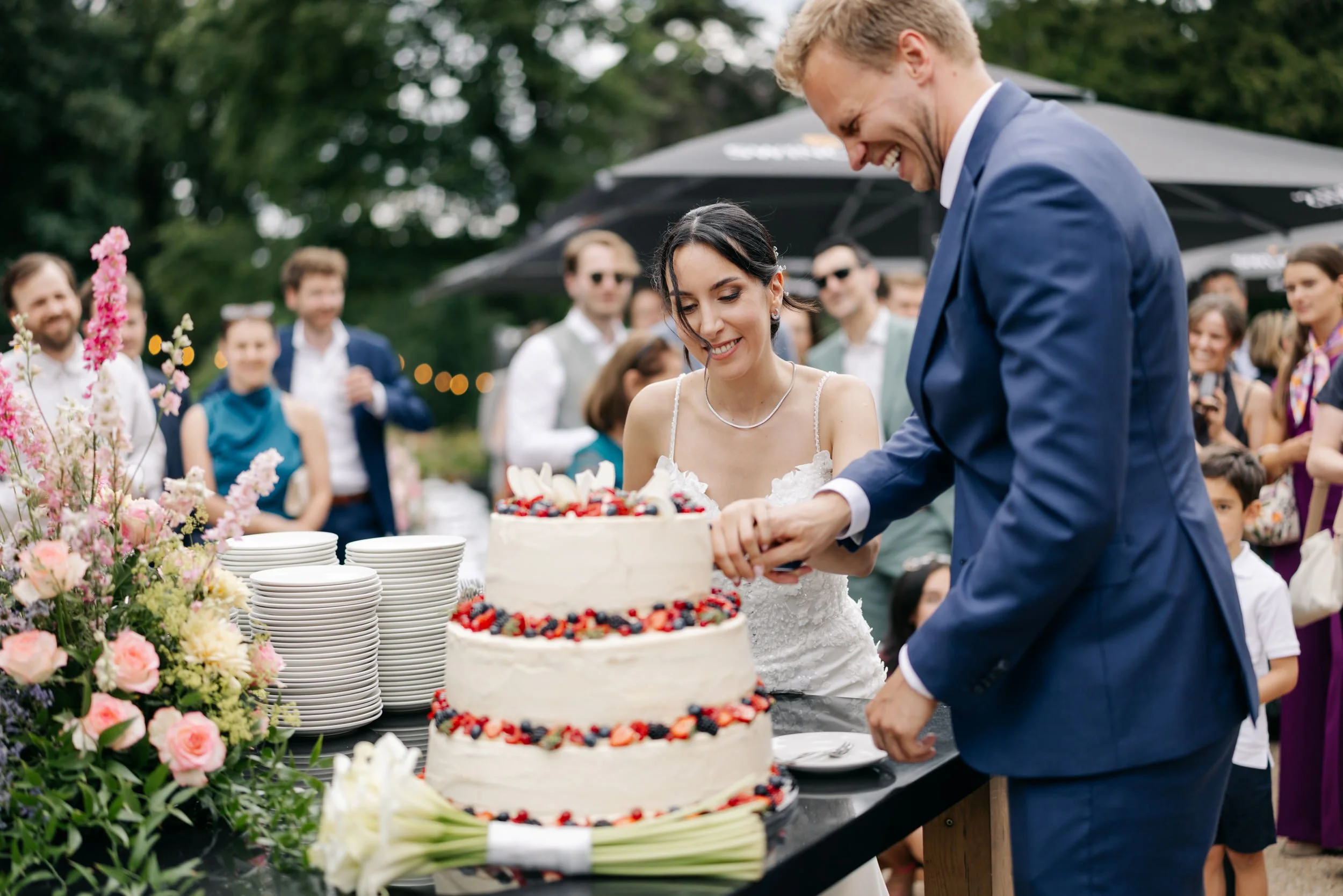  Bruidspaar snijdt de bruidstaart aan buiten, omringd door gasten en bloemen op tafel. 