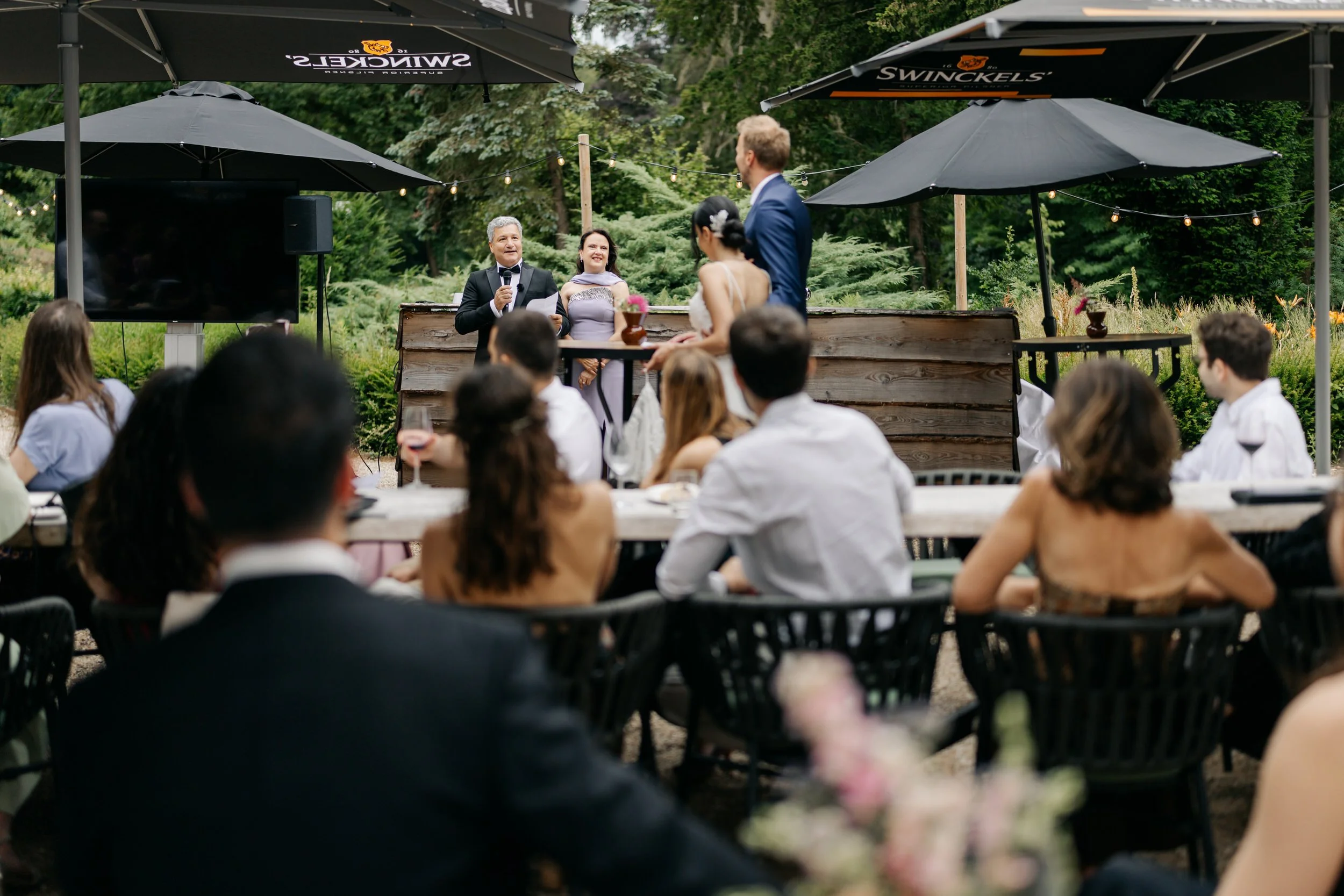  vader van de bruid houdt een speech op het terras terwijl de andere gasten aan lange tafels luisteren tijdens het diner buiten. 