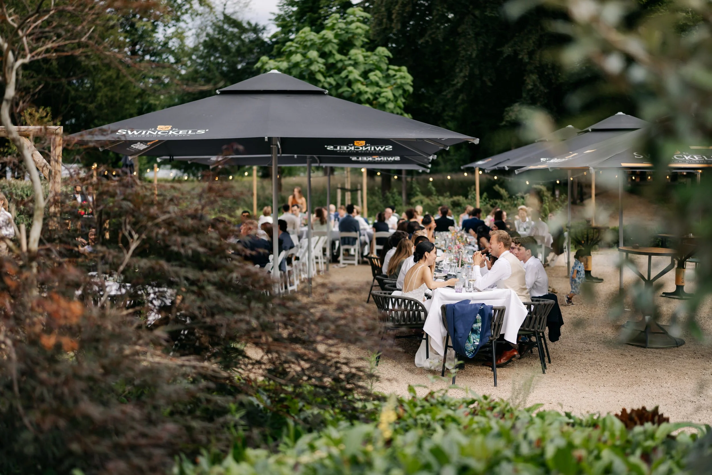  Gedekte lange tafels onder parasols op het terras, klaar voor het diner tijdens de bruiloft bij Jachtslot Mookerheide. 