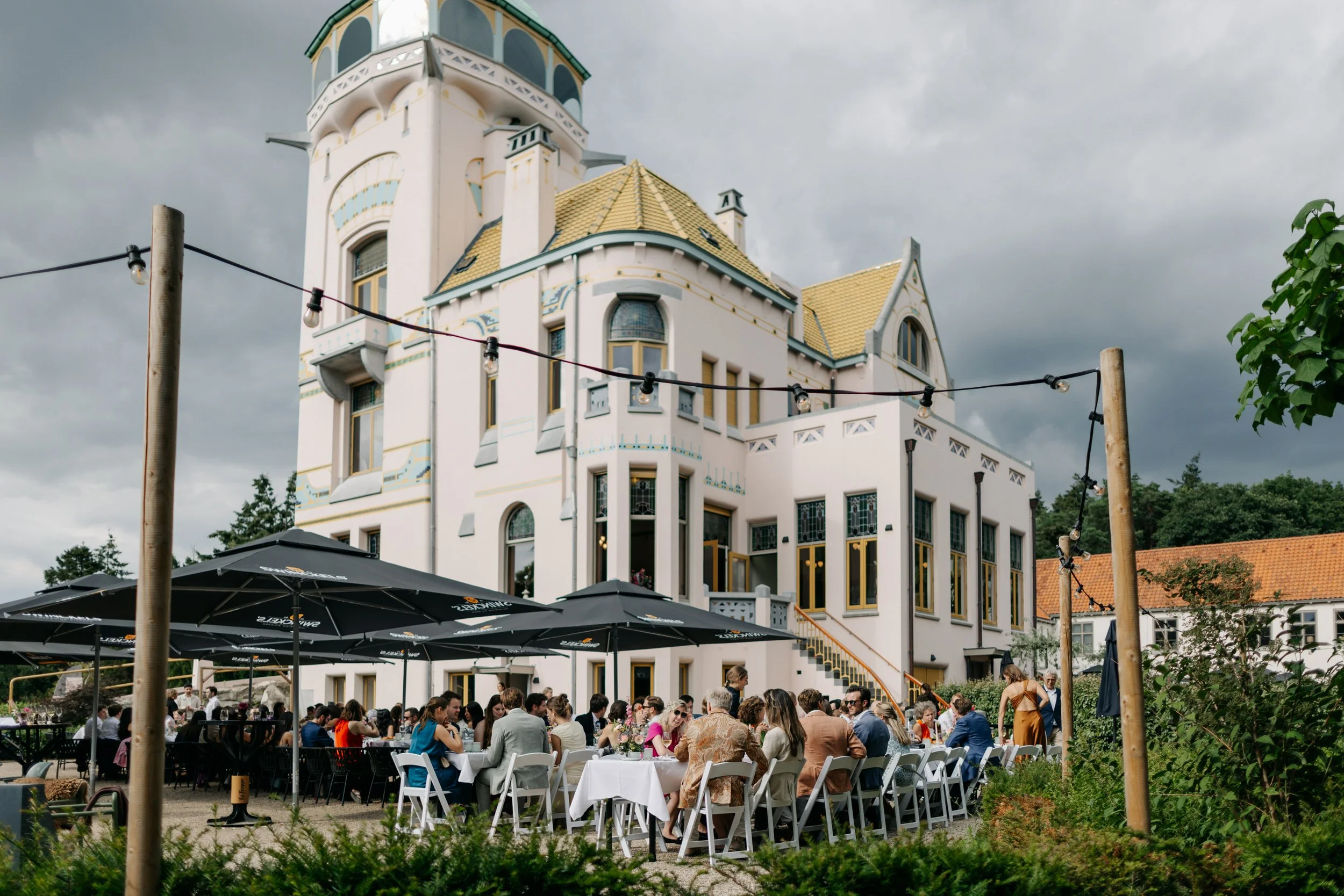 Internationale bruiloft bij jachtslot Mookerheide. Gasten zitten op het privé terras met op de achtergrond het jachtslot.