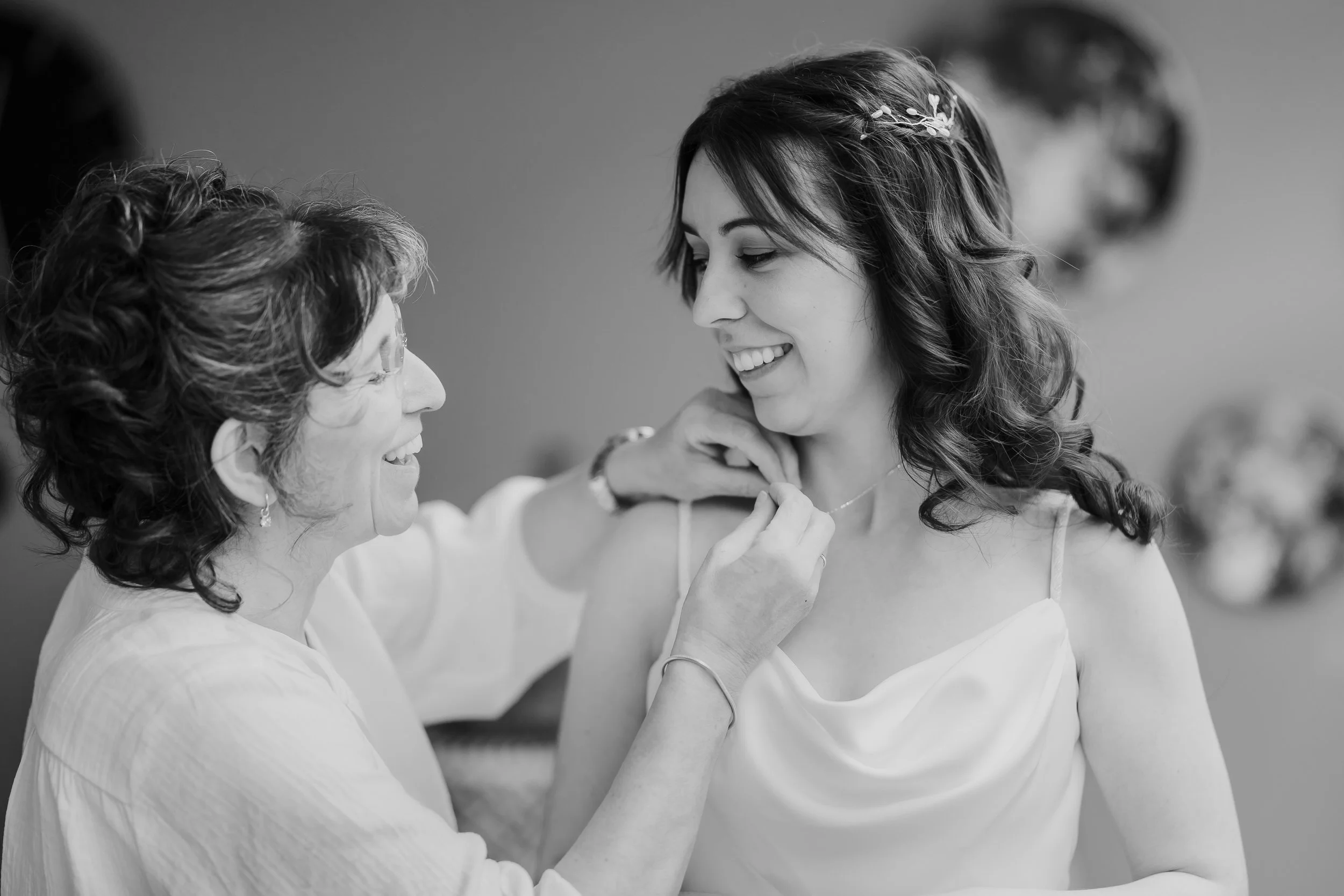 Bruid lacht tijdens de voorbereidingen terwijl ze geholpen wordt met haar ketting, zwart wit moment uit een trouwreportage