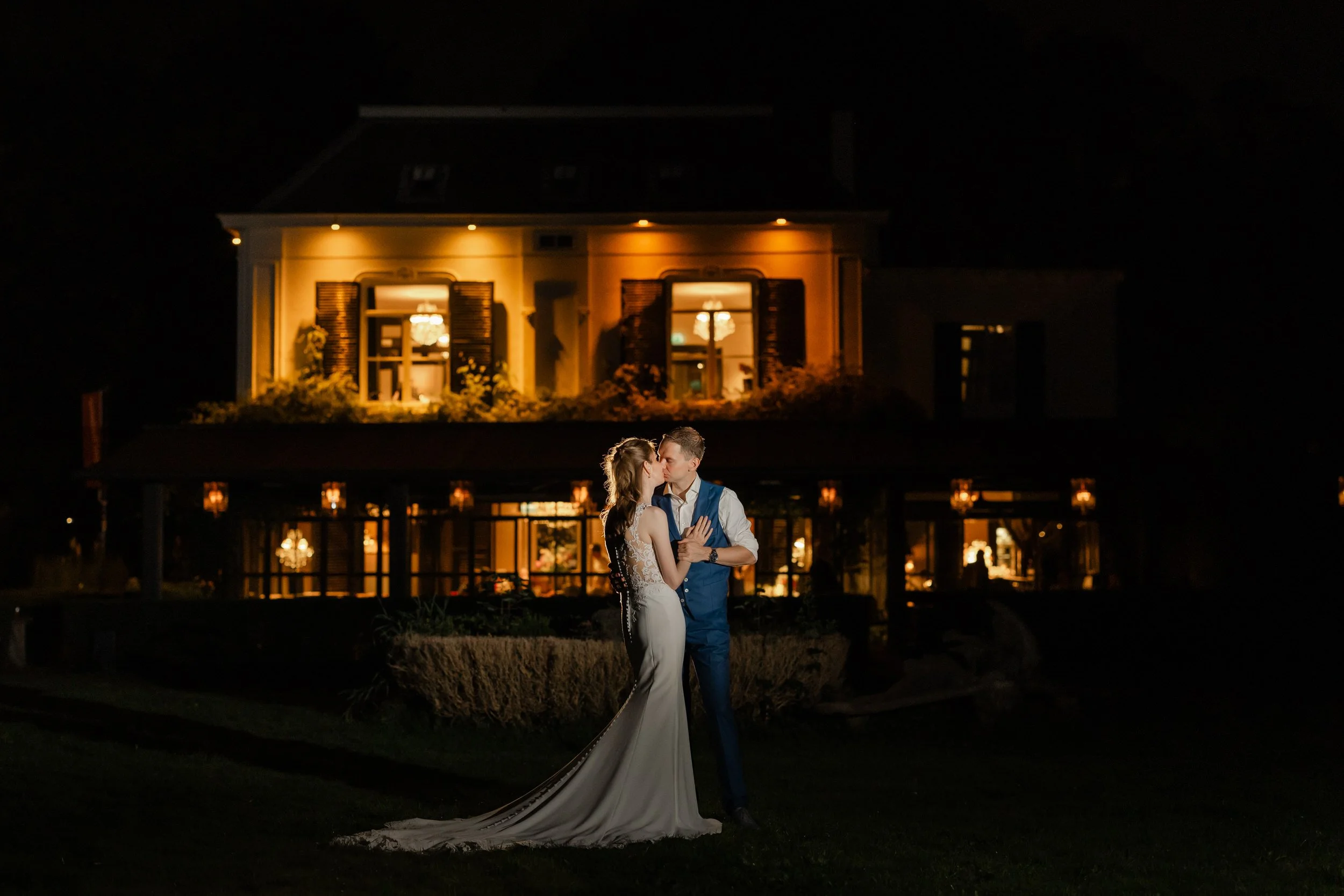 Bruidspaar staat samen voor een verlichte trouwlocatie in de avond, sfeervolle avondfoto tijdens de bruiloft bij landgoed Brakkesteyn.