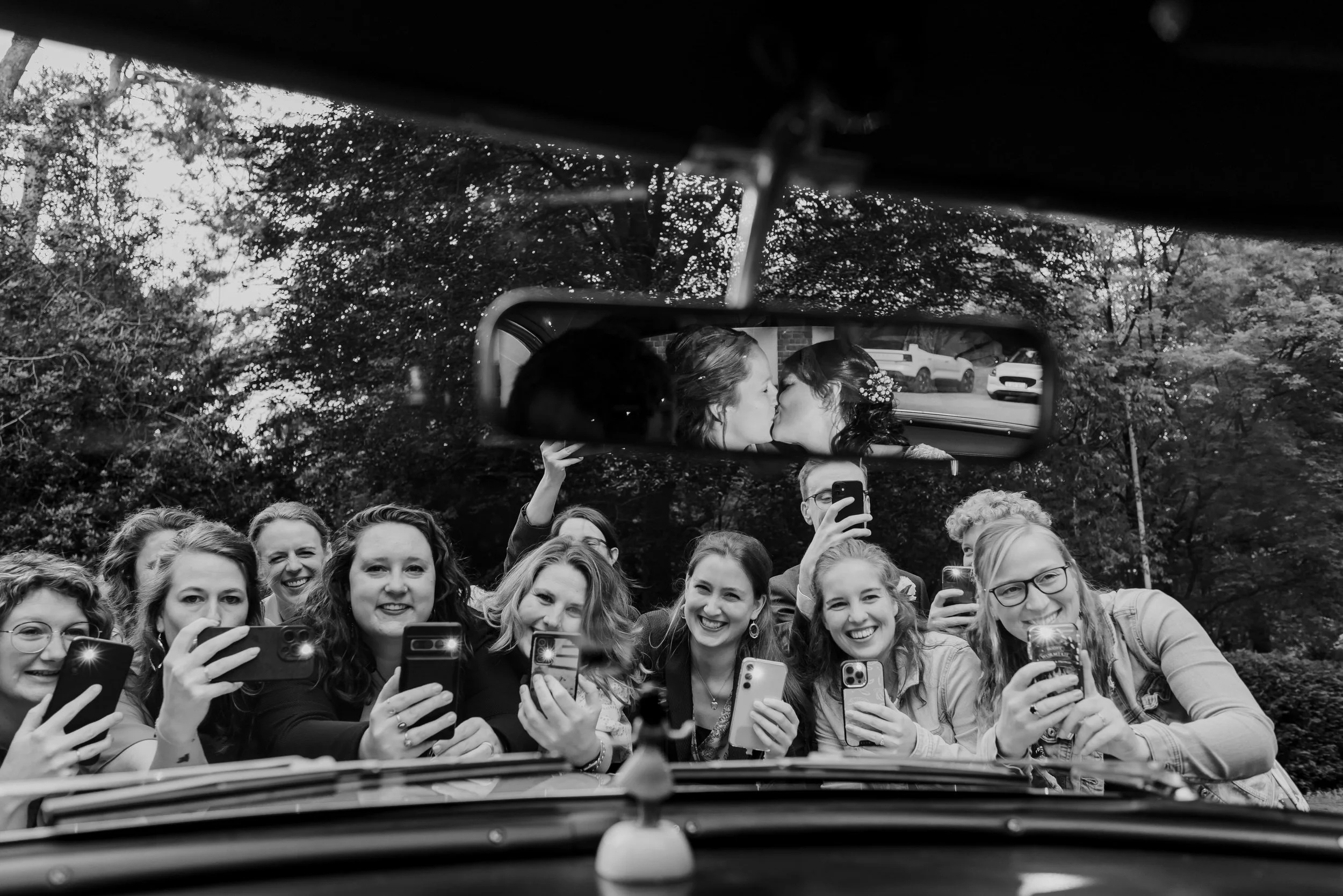 Bruidspaar kust in de auto terwijl gasten buiten enthousiast foto’s maken, zwart wit trouwfoto met een speelse editorial uitstraling