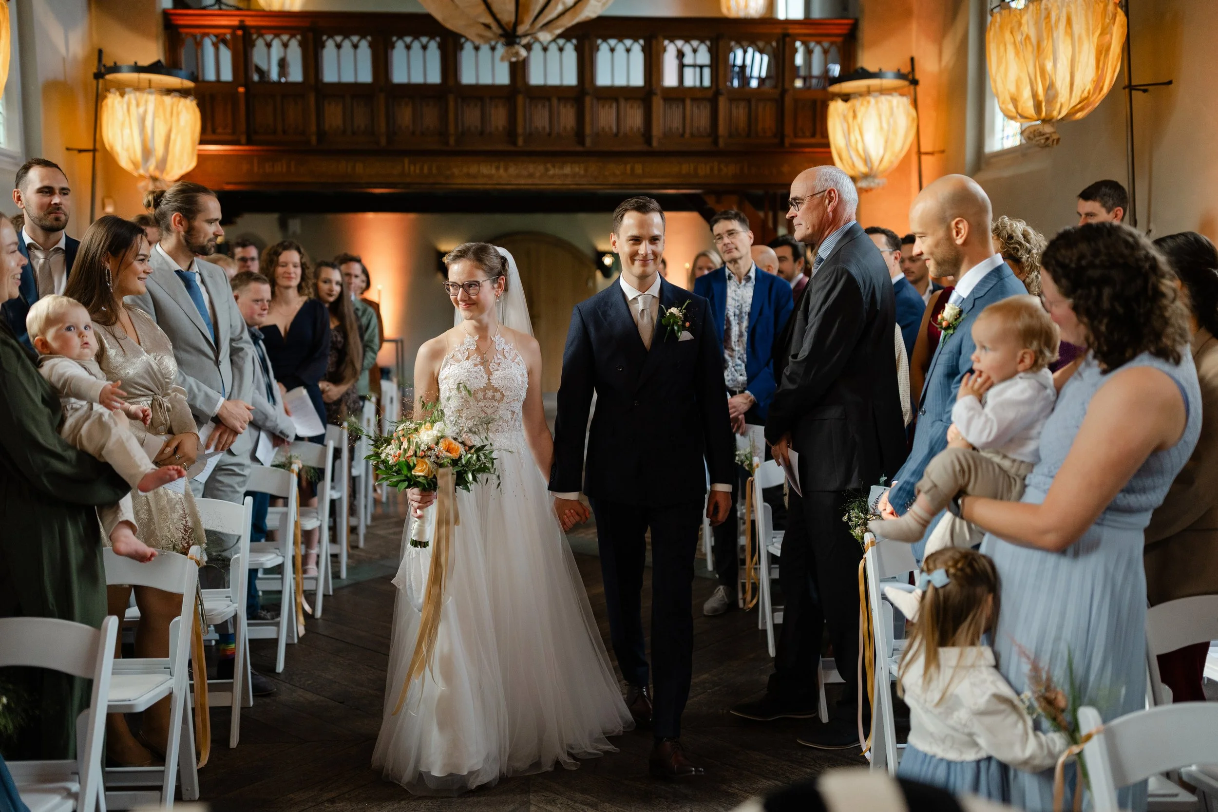 “Bruidspaar loopt hand in hand door het gangpad tijdens de ceremonie, omringd door gasten, gefotografeerd met een 35mm lens.”