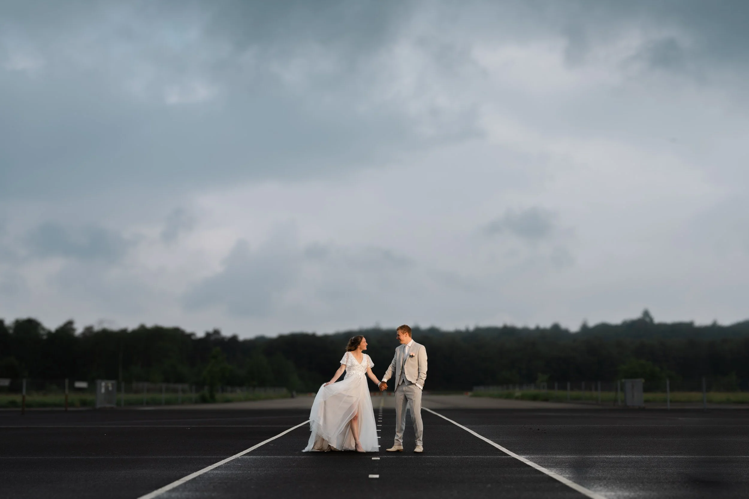 Bruidspaar staat samen op een lege weg onder een dreigende lucht, stijlvolle trouwfoto met veel ruimte en sfeer