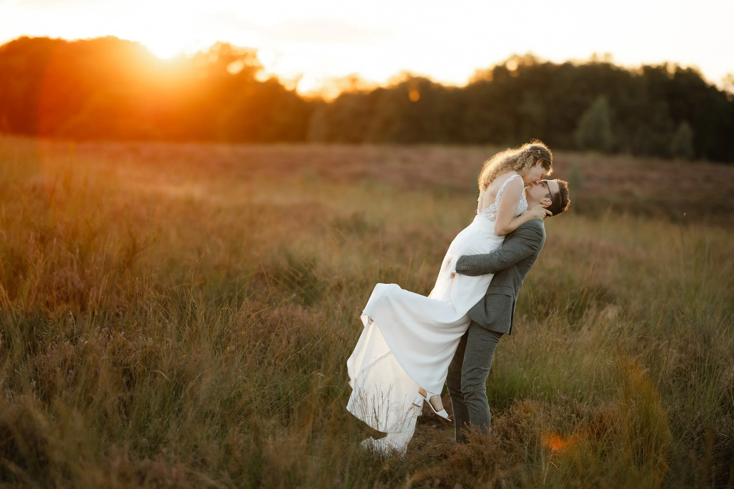 Bruidegom tilt de bruid op in een veld tijdens zonsondergang, romantische trouwfoto met warm avondlicht