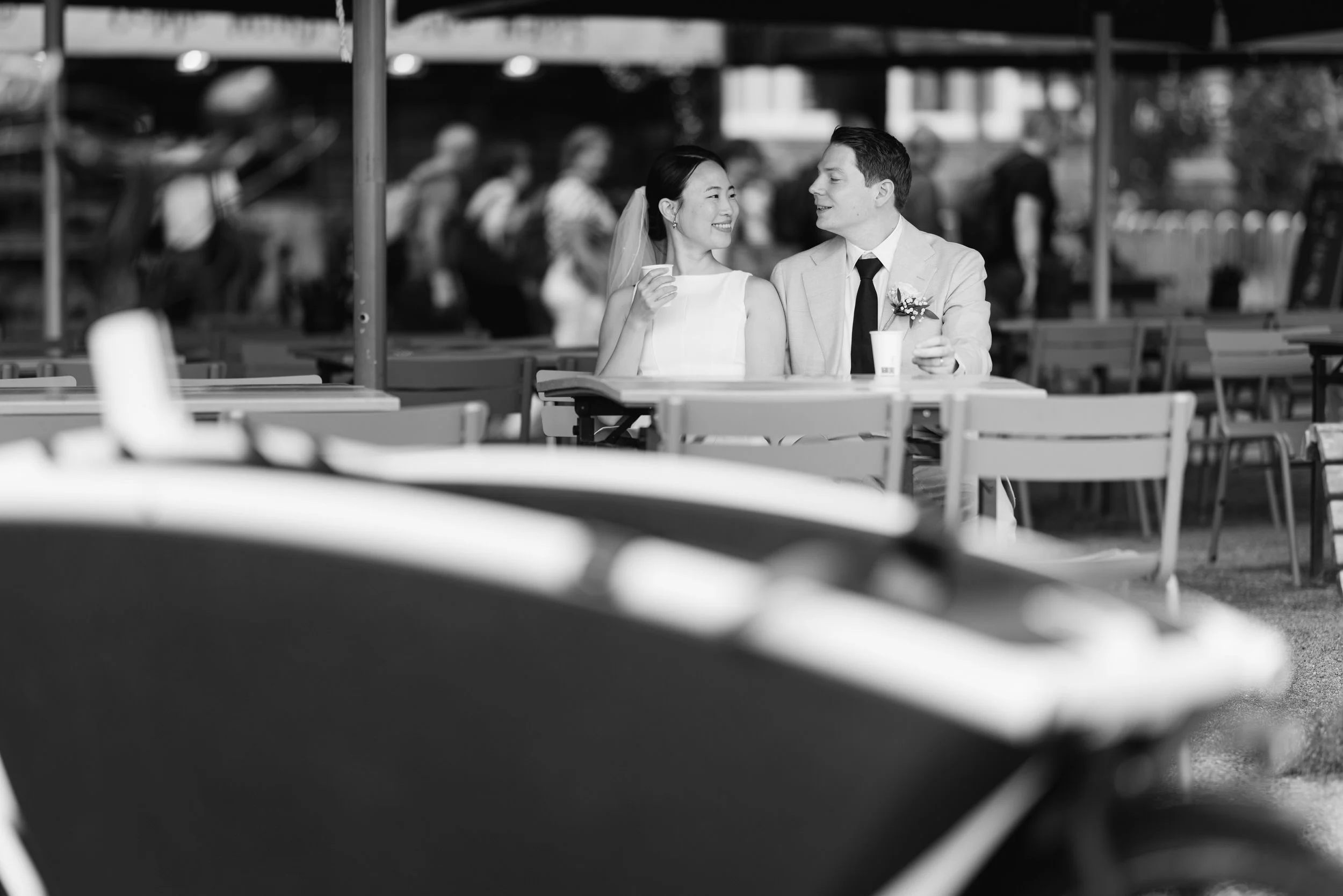 ruidspaar zit samen aan tafel en kijkt elkaar glimlachend aan tijdens een fotoshoot in Amsterdam, zwart wit trouwfoto met rustige sfeer