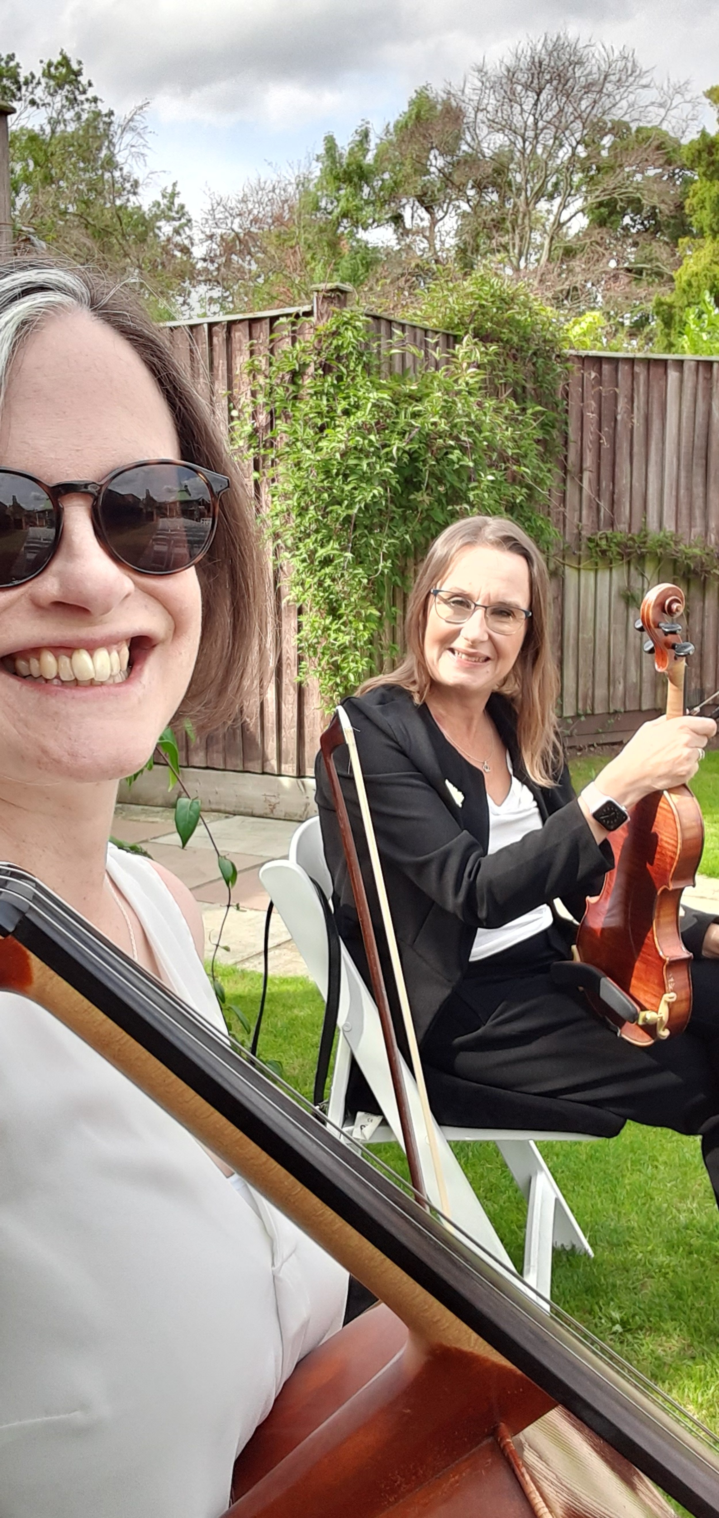 Two women outdoors, one smiling with sunglasses holding a guitar, the other with a serious expression playing a violin, in a backyard with a wooden fence and trees.