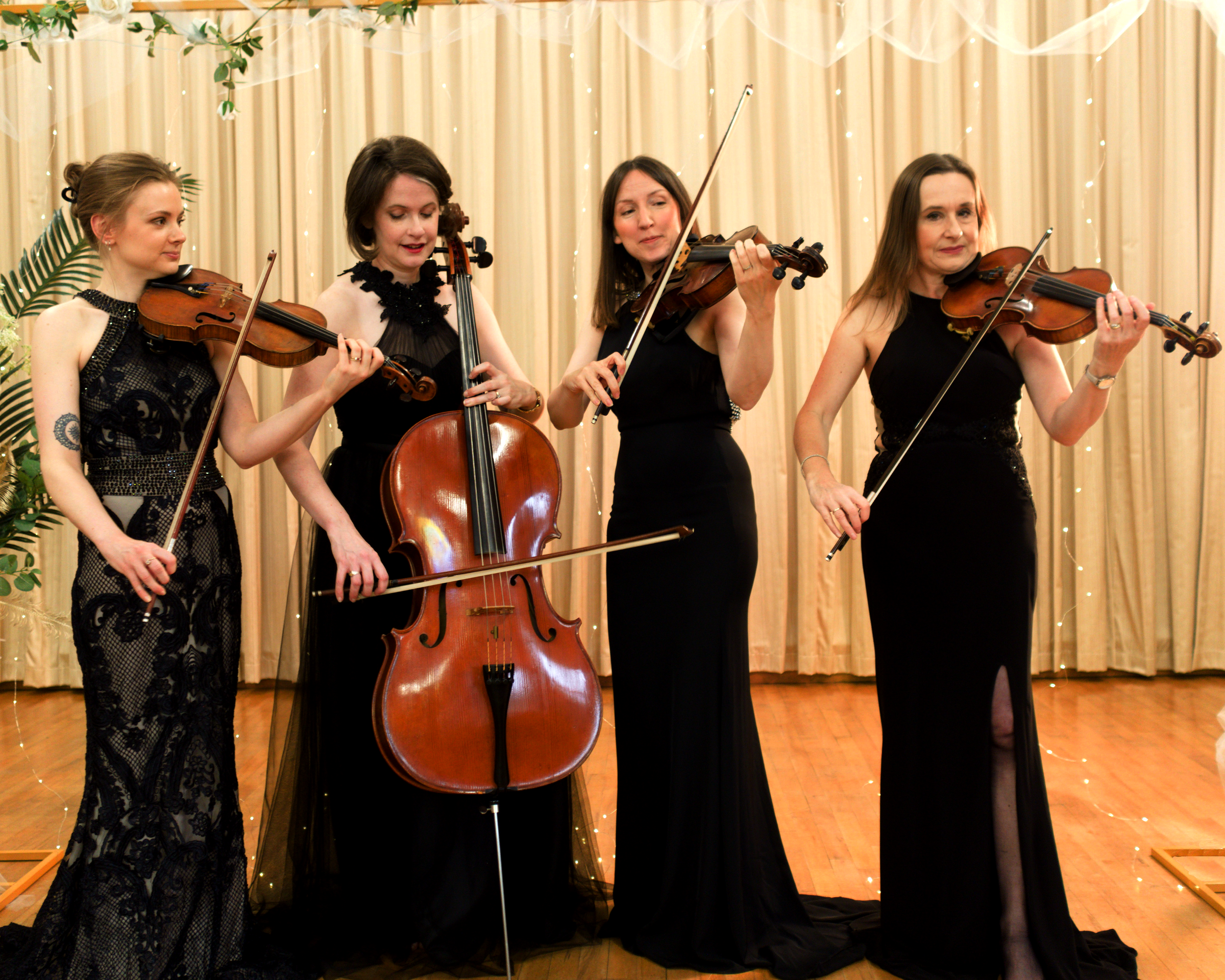 Four women in formal dresses playing string instruments on stage, with cream-colored curtains and string lights in the background.