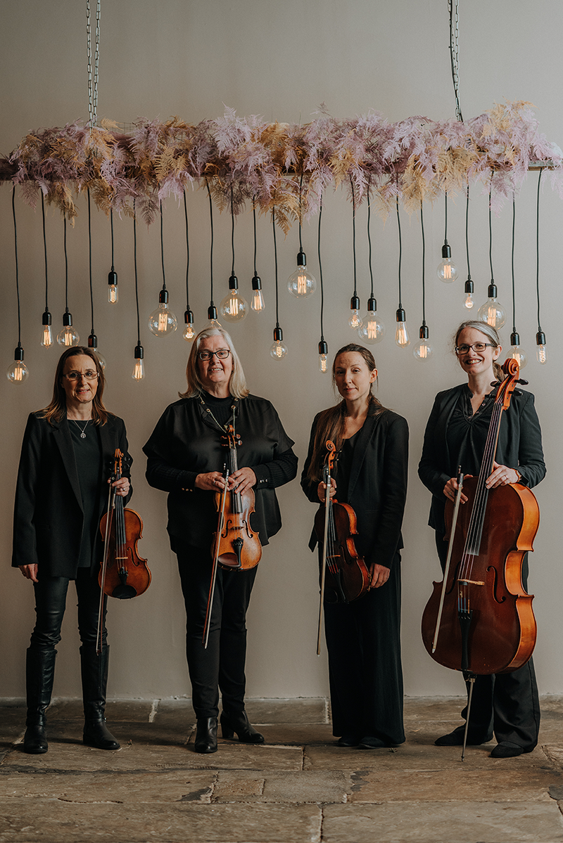 The Fern Quartet stand in The Arches, Dean Clough, against white wall and under an arrangement of features and hanging light bulbs