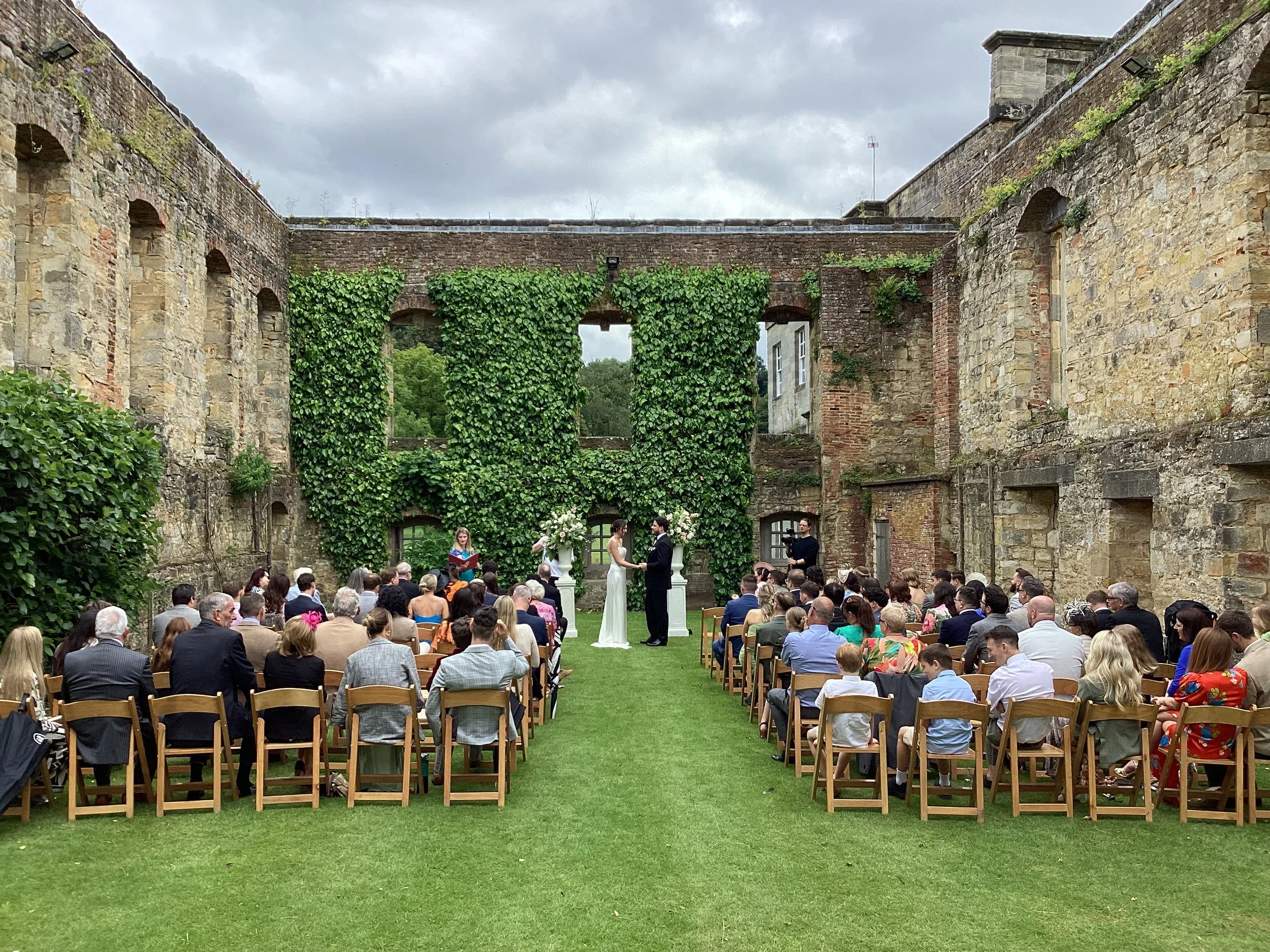 Guests sat at an outdoor wedding, in the ruins of Newburgh Priory, surrounded by greenary. The couple at the front standing facing each other and holding hands, the celebrant off to the left
