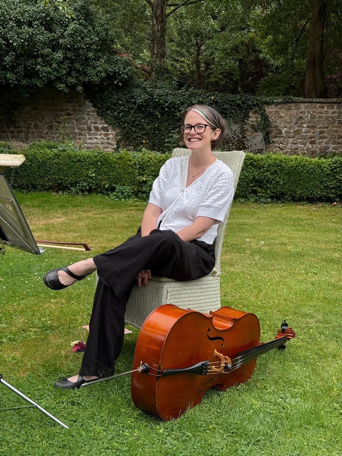 Woman sitting outdoors on a chair with a cello lying on the grass nearby, smiling with crossed legs, in a garden setting with trees and a stone wall in the background.