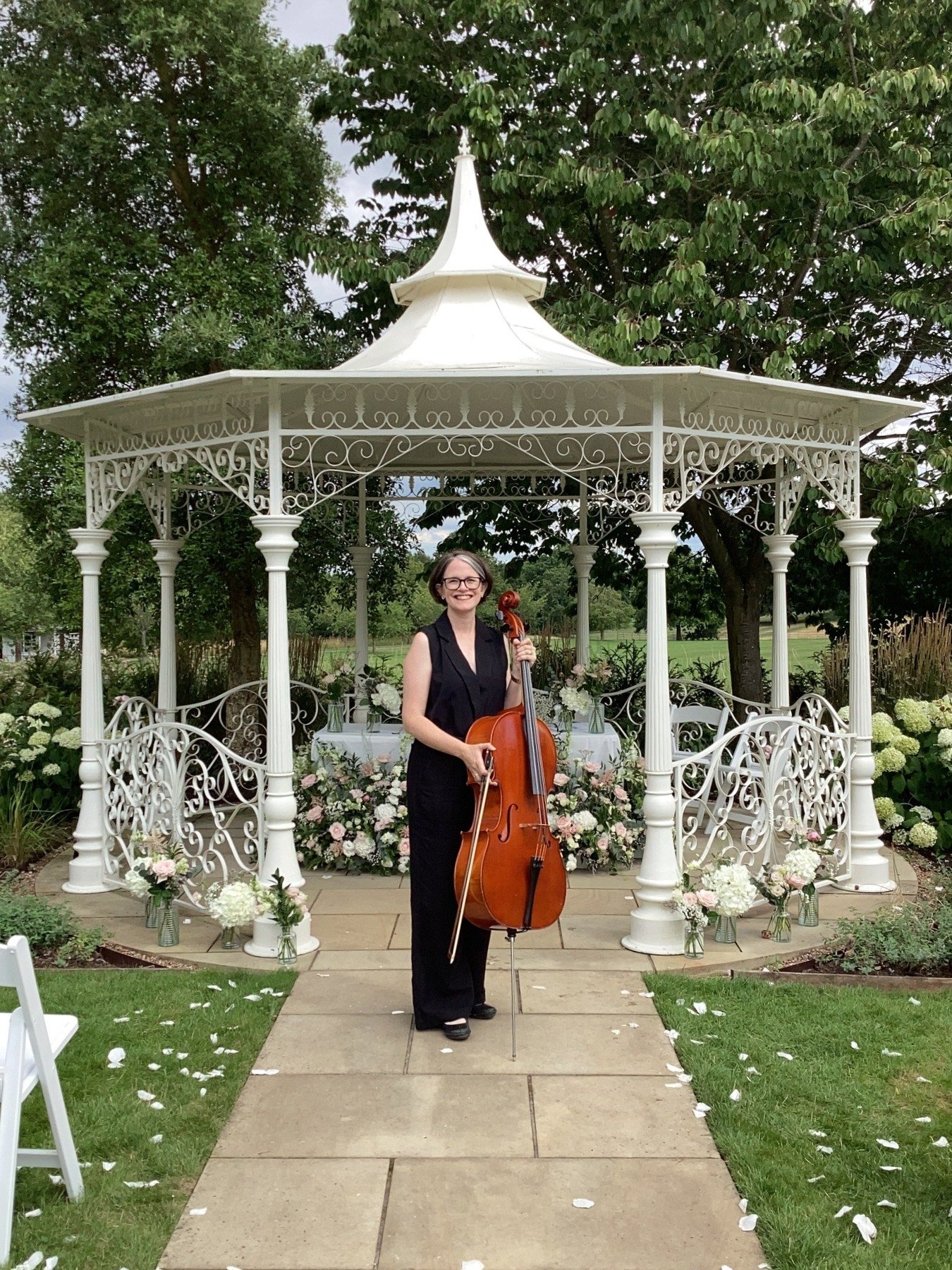 Cellist stands infront of the white gazibo in the grounds of Norwood Park, surrounded by white confetti petals