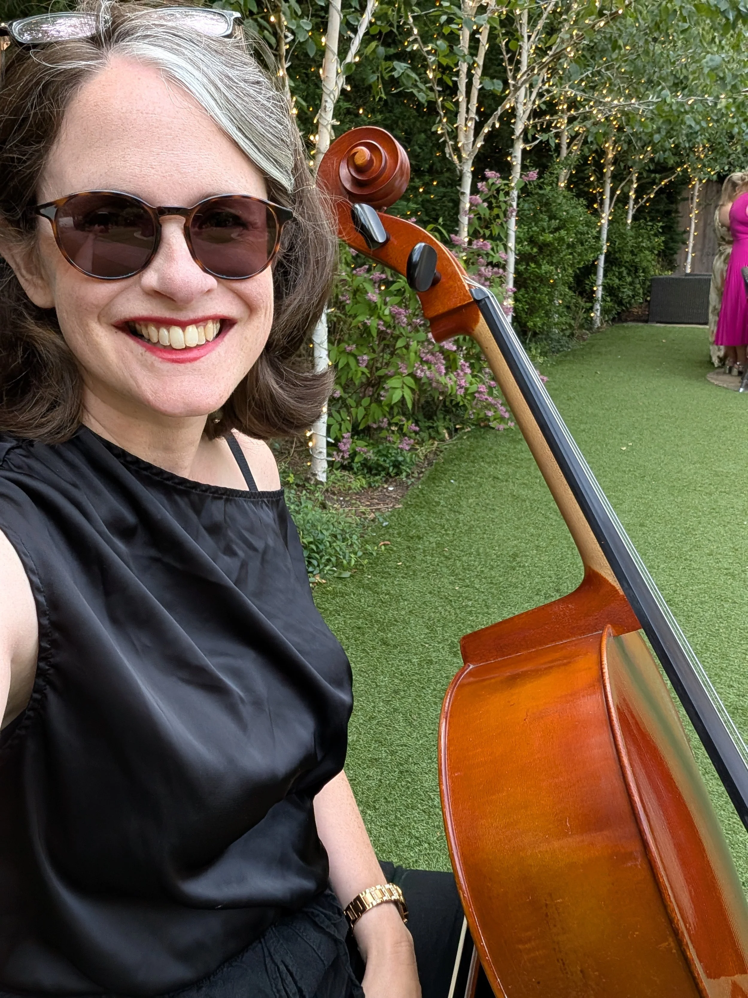 A woman with gray hair and sunglasses smiling, wearing a black sleeveless top, sitting outdoors near a string-light decorated garden with trees and bushes. A cello is visible next to her.