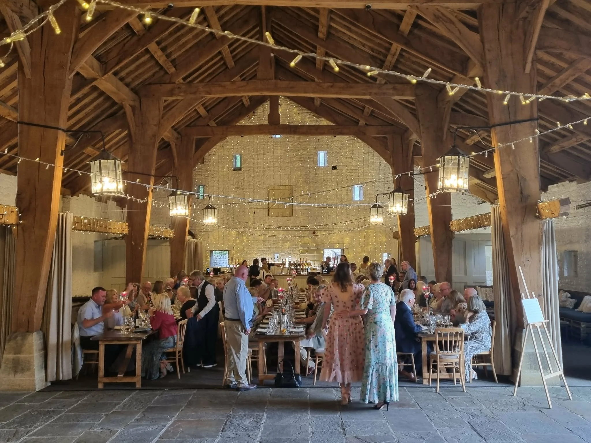 Interior of airedale barn set up for a reception with people seated. Fairy lights hange across wooden beams over head and lanterns are light above each table