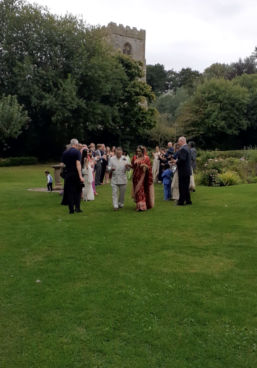 A wedding ceremony taking place outdoors on a grassy lawn, with a large group of people dressed in formal attire, and a historic stone tower and trees in the background.