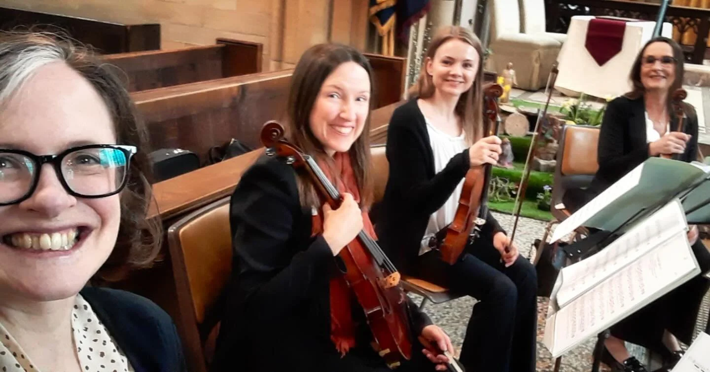 Four women sitting in an indoor setting, two holding violins, smiling at the camera during a musical practice or rehearsal.
