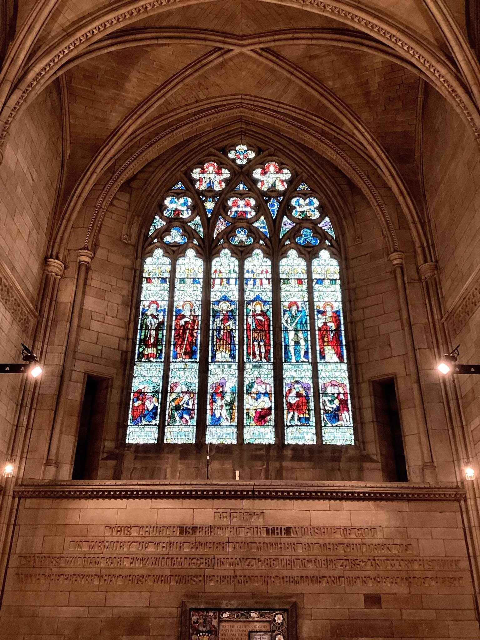 A large stained glass window inside a church depicting religious figures and scenes, with intricate stonework and a pointed arch design.