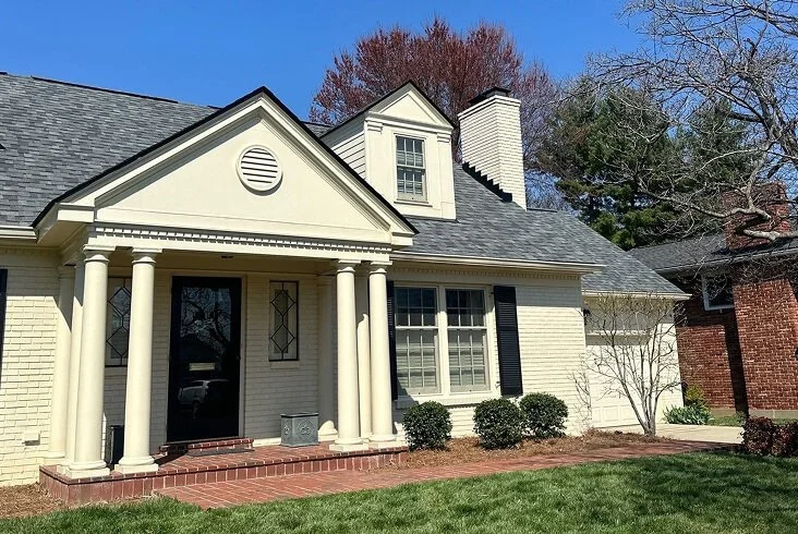 Completed gray shingle roof on a colonial-style cream painted brick home with columned front porch, black shutters, and a dormer window