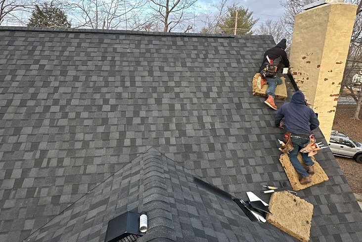 Roofing crew installing dark gray architectural shingles near the ridge of a roof next to a chimney