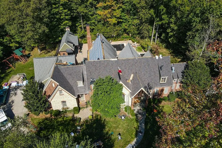 Aerial view of a roofing crew replacing shingles on a large brick and stone home with a complex roofline surrounded by mature trees