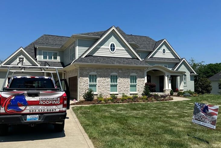 Completed dark gray shingle roof on a craftsman-style home with stone and siding exterior, with a Unisource Roofing truck and yard sign in the driveway