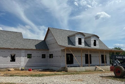 New gray shingle roof installed on a large home under construction with house wrap on the exterior walls, arched dormers, and a covered front porch framed in wood