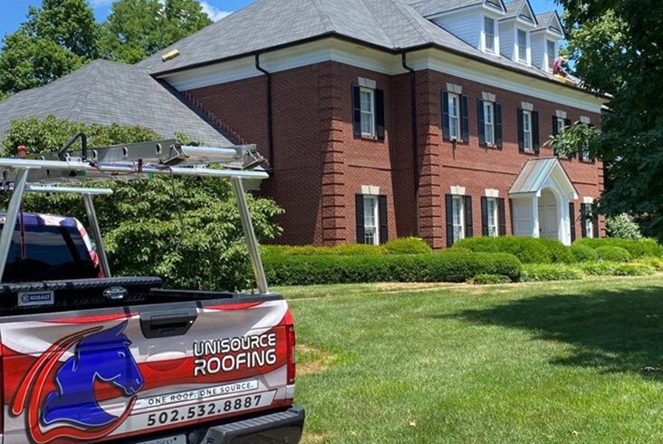 Unisource Roofing truck parked in front of a large two-story red brick home with manicured hedges while crew members work on the roof