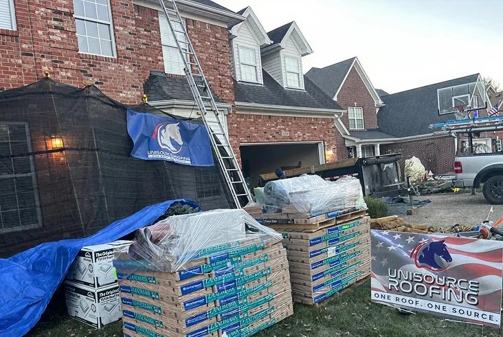 Pallets of bundled roofing shingles stacked in the driveway of a brick two-story home with a Unisource Roofing banner and ladder leaning against the house