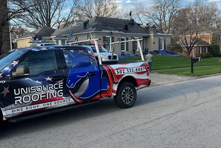 Unisource Roofing branded truck parked on the street while a crew works on a residential roof replacement in the background with blue tarps on the lawn