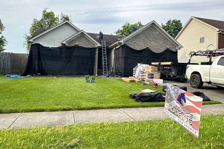 Roofing crew working on a single-story home with protective black tarps hung along the exterior, shingle bundles on the ground, and a Unisource Roofing yard sign and work truck in view