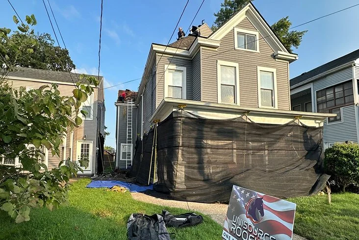 Roofing crew replacing the roof on a two-story Victorian-style home with protective mesh tarps around the lower level and a Unisource Roofing yard sign on the lawn