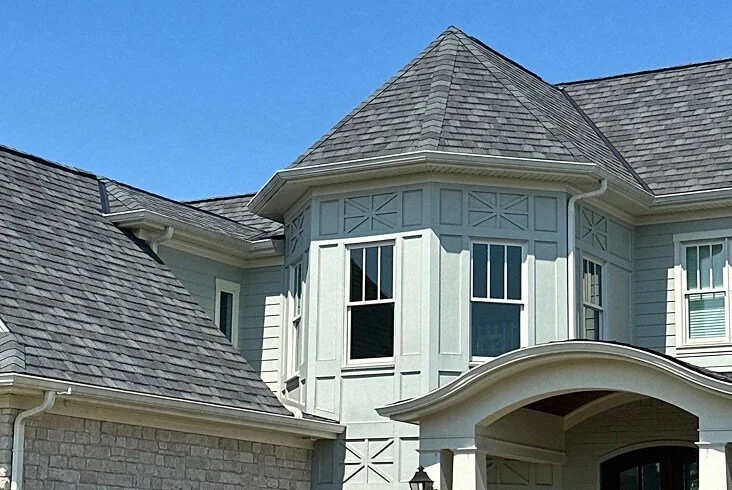 Gray architectural shingles on a multi-angled roofline featuring a turret-style bay window with decorative trim on a light green home