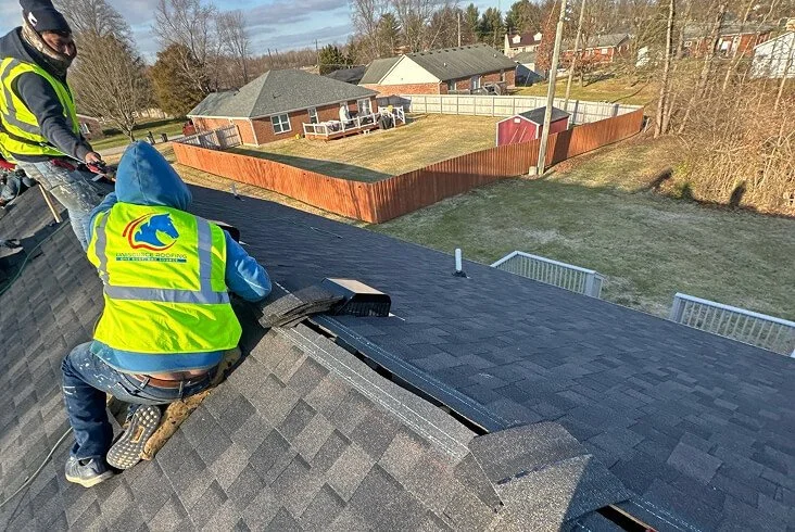 Unisource Roofing crew members in branded safety vests installing dark shingles on a residential roof overlooking a suburban backyard