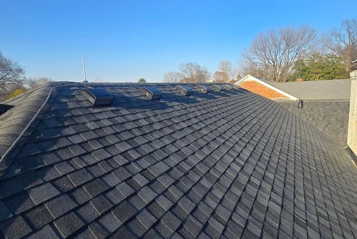 Completed dark gray asphalt shingle roof with a row of metal roof vents under a clear blue sky