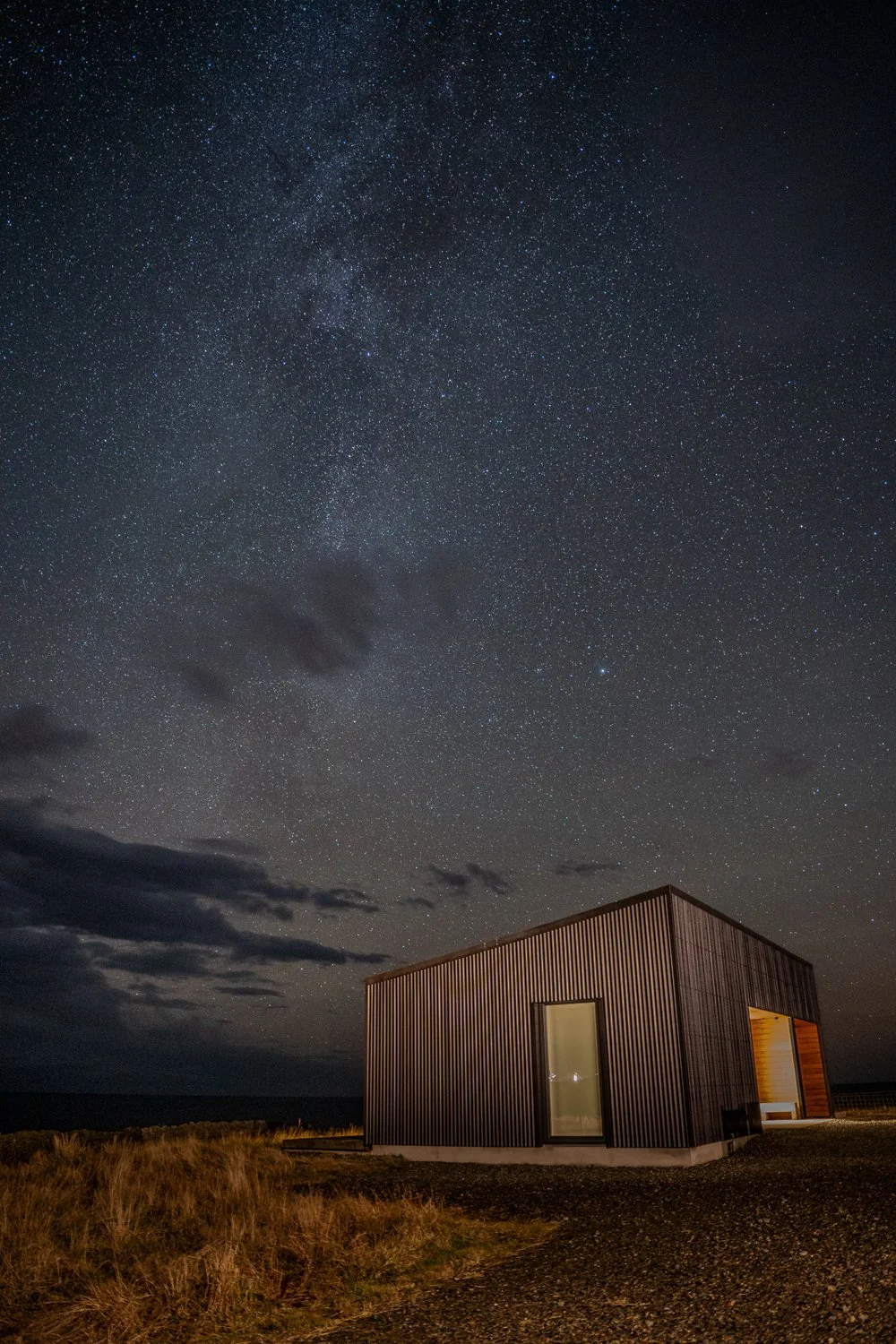 Brighid illuminated at night beneath a stunning starry sky. The warm glow of the cottage contrasts beautifully with the dark, expansive heavens, creating a serene and magical atmosphere. 