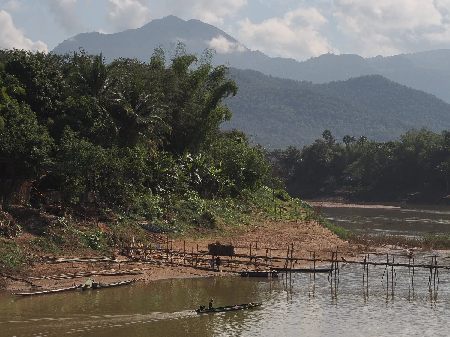 A man on a fishing boat on a river in Luang Prabang, with mountains in the background.